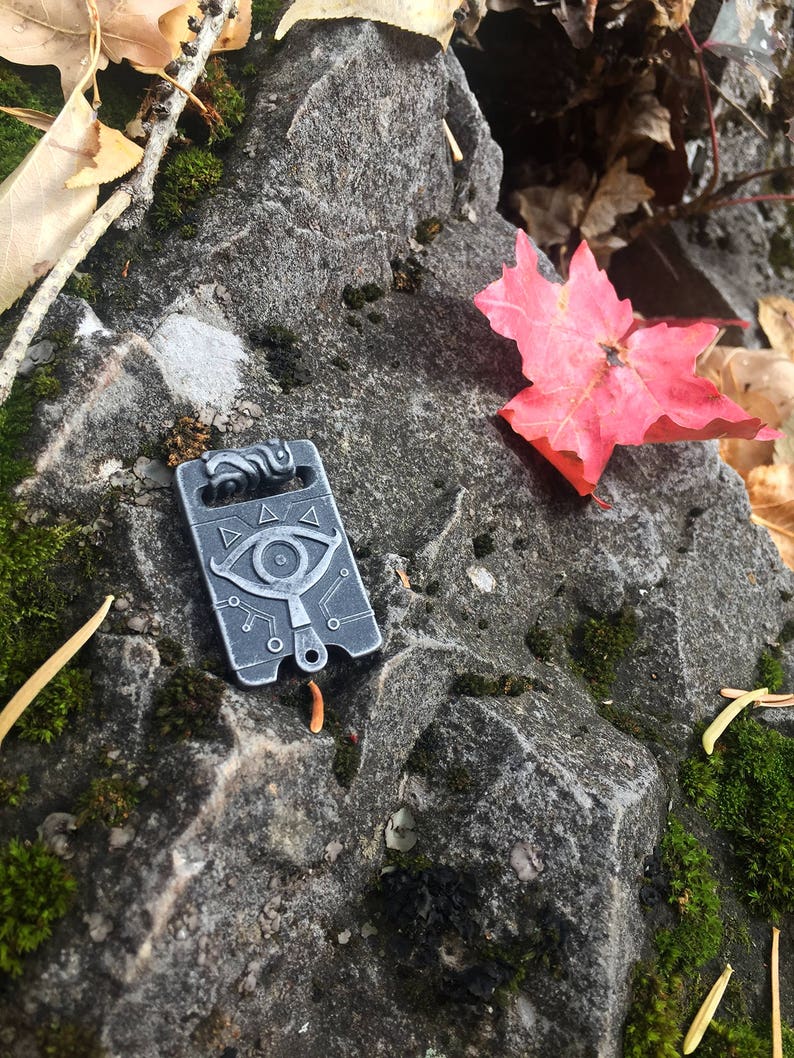 May include: A silver metal charm with a stylised eye design and geometric patterns. The charm is on a rocky surface with a red maple leaf.