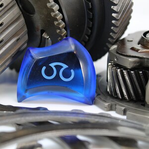 May include: A close-up of a blue plastic gear shift knob with a white symbol. The knob is surrounded by metal gears.