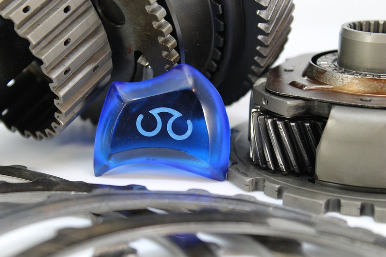 May include: A close-up of a blue plastic gear shift knob with a white symbol. The knob is surrounded by metal gears.