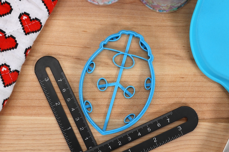 May include: A blue plastic ladybug cookie cutter. The cutter is shaped like a ladybug with a round body and six legs. There are five small circles on the body of the ladybug.