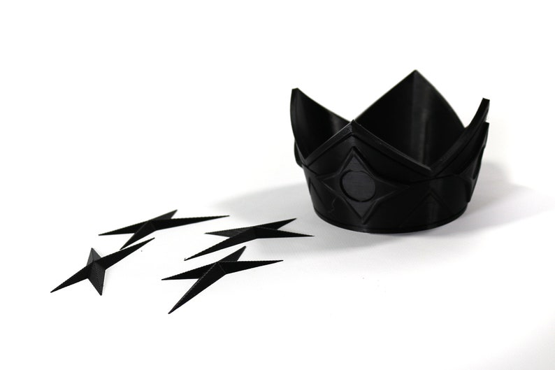 May include: A black plastic crown with a geometric design and a black circle in the center. Five black plastic stars are scattered around the crown.