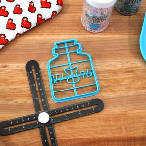 May include: Blue plastic cookie cutter shaped like a jar with the word "Honey" written on it. The cutter is on a wooden surface with a black ruler and a blue spatula.