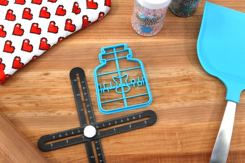 May include: Blue plastic cookie cutter shaped like a jar with the word "Honey" written on it. The cutter is on a wooden surface with a black ruler and a blue spatula.