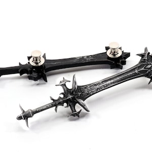 May include: Two black metal sword pins with silver accents. The swords have intricate details and are attached to a pin back.