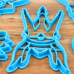 May include: A collection of light blue plastic cookie cutters in various shapes, including a crown, a lightning bolt, and other abstract designs. The cutters are arranged on a light brown wooden surface.
