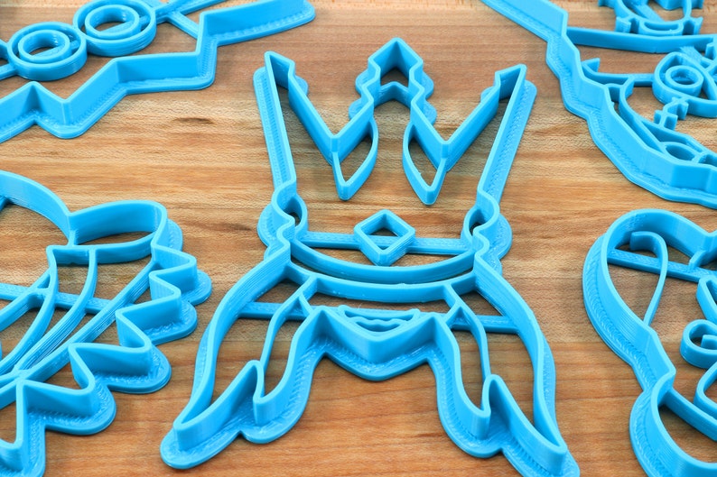 May include: A collection of light blue plastic cookie cutters in various shapes, including a crown, a lightning bolt, and other abstract designs. The cutters are arranged on a light brown wooden surface.