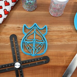 May include: A blue plastic cookie cutter shaped like a cartoon monster with a crown, horns, and a wide open mouth. The cookie cutter is on a wooden surface with a ruler, rolling pin, and spatula.