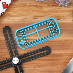 May include: A blue plastic cookie cutter shaped like a handheld video game console. The cutter is on a wooden surface with a black ruler and a blue spatula.