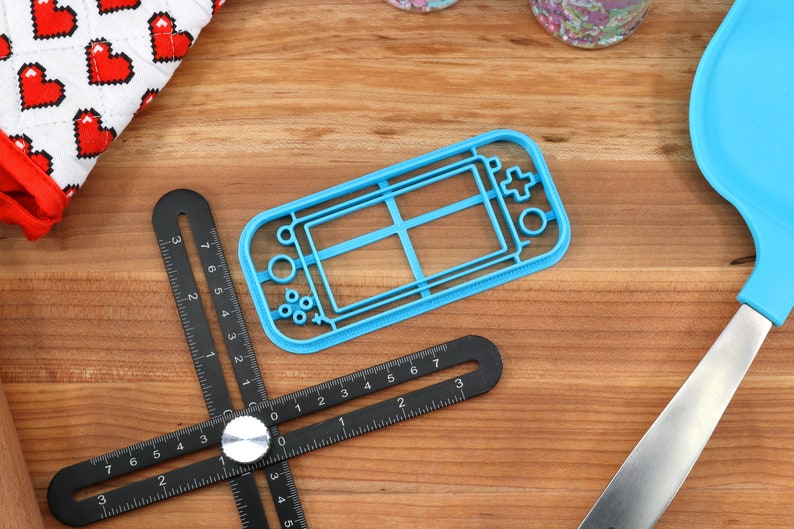May include: A blue plastic cookie cutter shaped like a handheld video game console. The cutter is on a wooden surface with a black ruler and a blue spatula.