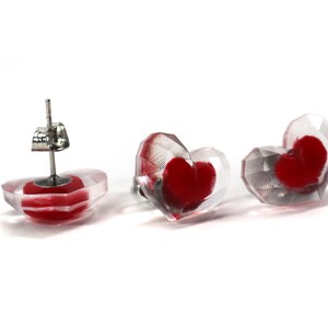 Puede incluir: Un par de pendientes en forma de coraz&oacute;n con una base de acr&iacute;lico transparente y un coraz&oacute;n rojo en el interior. Los pendientes tienen un poste de plata y una mariposa en la parte posterior.