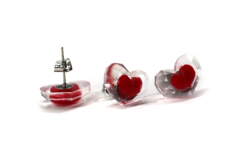Puede incluir: Un par de pendientes en forma de coraz&oacute;n con una base de acr&iacute;lico transparente y un coraz&oacute;n rojo en el interior. Los pendientes tienen un poste de plata y una mariposa en la parte posterior.