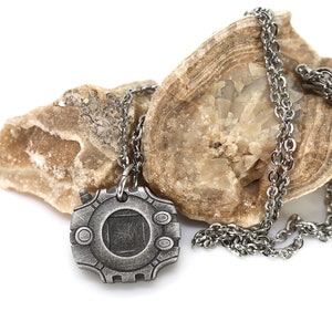 Puede incluir: Un collar de plata con un colgante con forma de dispositivo digital. El colgante tiene una pantalla cuadrada y un bot&oacute;n circular a cada lado. El collar est&aacute; apoyado en una roca marr&oacute;n.
