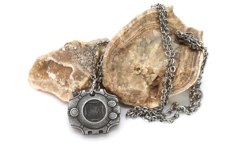 Puede incluir: Un collar de plata con un colgante con forma de dispositivo digital. El colgante tiene una pantalla cuadrada y un bot&oacute;n circular a cada lado. El collar est&aacute; apoyado en una roca marr&oacute;n.