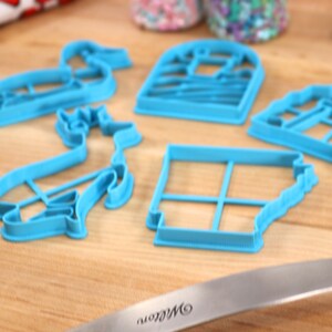Op de afbeelding: Vijf blauwe plastic koekjesvormen in verschillende vormen, waaronder een eend, een hert, een huis, een windmolen en een vierkant met een rasterpatroon. De vormen liggen op een houten oppervlak.
