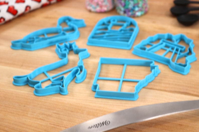 Op de afbeelding: Vijf blauwe plastic koekjesvormen in verschillende vormen, waaronder een eend, een hert, een huis, een windmolen en een vierkant met een rasterpatroon. De vormen liggen op een houten oppervlak.