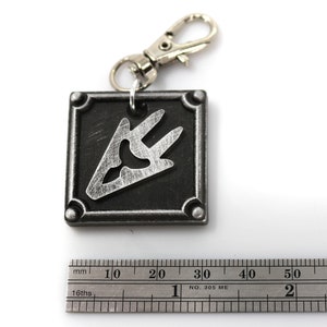 May include: A silver metal keychain with a black square charm featuring a silver stylized symbol. The charm has a slightly textured surface.