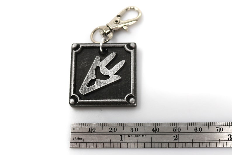 May include: A silver metal keychain with a black square charm featuring a silver stylized symbol. The charm has a slightly textured surface.