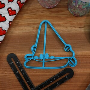 May include: A blue plastic cookie cutter shaped like a sailboat. The cutter is on a wooden surface.