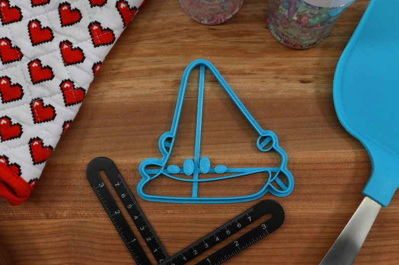May include: A blue plastic cookie cutter shaped like a sailboat. The cutter is on a wooden surface.