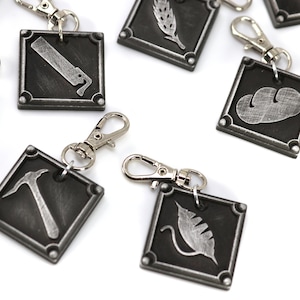 May include: A set of eight silver keychains with different symbols, including a hammer, a leaf, a wheat stalk, a cloud, and a knife. Each keychain has a silver clip and a square-shaped tag with a black background.