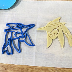 May include: Two cookie cutters, one blue and one yellow, shaped like a person wearing a hat. The cutters are on a white piece of parchment paper.