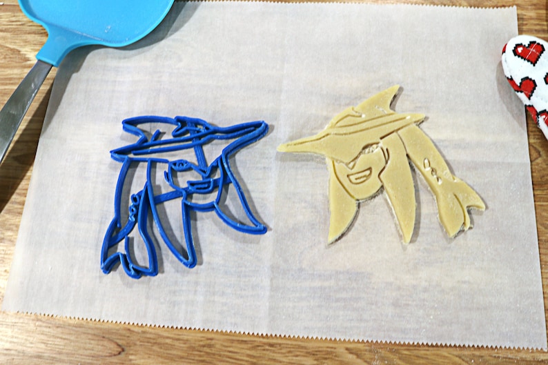 May include: Two cookie cutters, one blue and one yellow, shaped like a person wearing a hat. The cutters are on a white piece of parchment paper.
