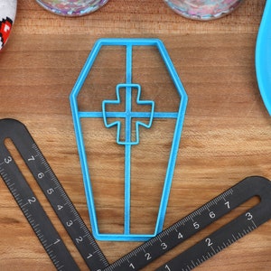 May include: A blue plastic cookie cutter shaped like a coffin with a cross inside. The cutter is on a wooden surface.