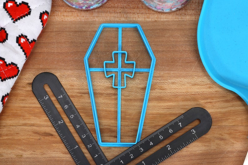 May include: A blue plastic cookie cutter shaped like a coffin with a cross inside. The cutter is on a wooden surface.