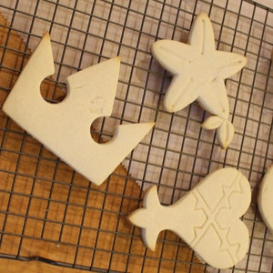 Op de afbeelding: Zes witte suikerkoekjes op een draadrek. De koekjes zijn gevormd als een kroon, een zeester, een hart, een cirkel en twee verschillende abstracte vormen.
