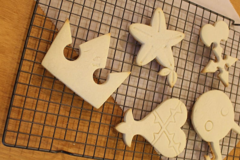 Op de afbeelding: Zes witte suikerkoekjes op een draadrek. De koekjes zijn gevormd als een kroon, een zeester, een hart, een cirkel en twee verschillende abstracte vormen.