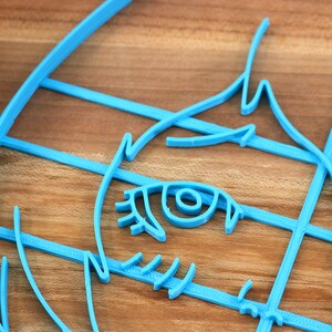 May include: A blue plastic cookie cutter shaped like a cartoon face with one eye visible. The cutter is on a wooden surface.