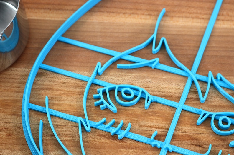 May include: A blue plastic cookie cutter shaped like a cartoon face with one eye visible. The cutter is on a wooden surface.
