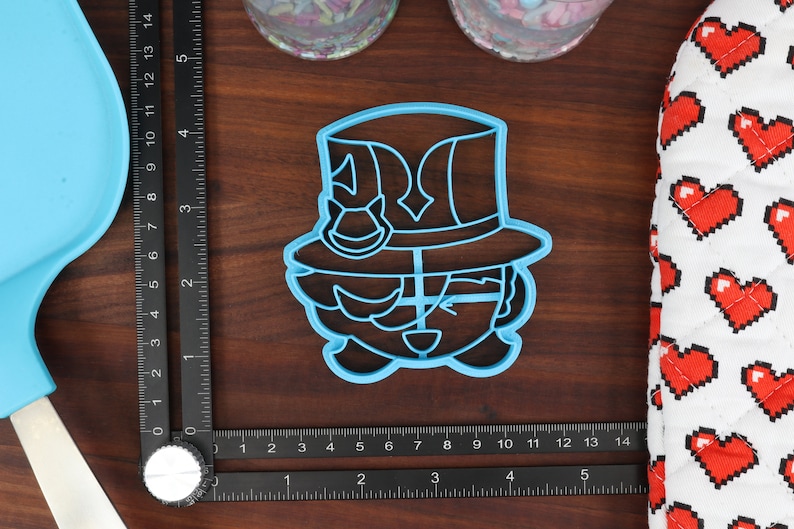 May include: A blue plastic cookie cutter shaped like a cartoon character wearing a hat. The character has a large hat with a brim and a small, round face with a smile.