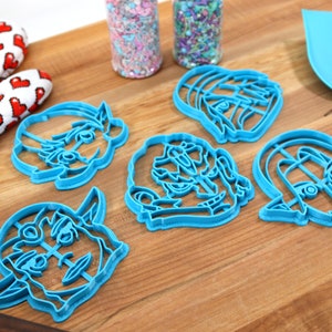 May include: Five blue plastic cookie cutters shaped like cartoon faces. The cutters are on a wooden surface.