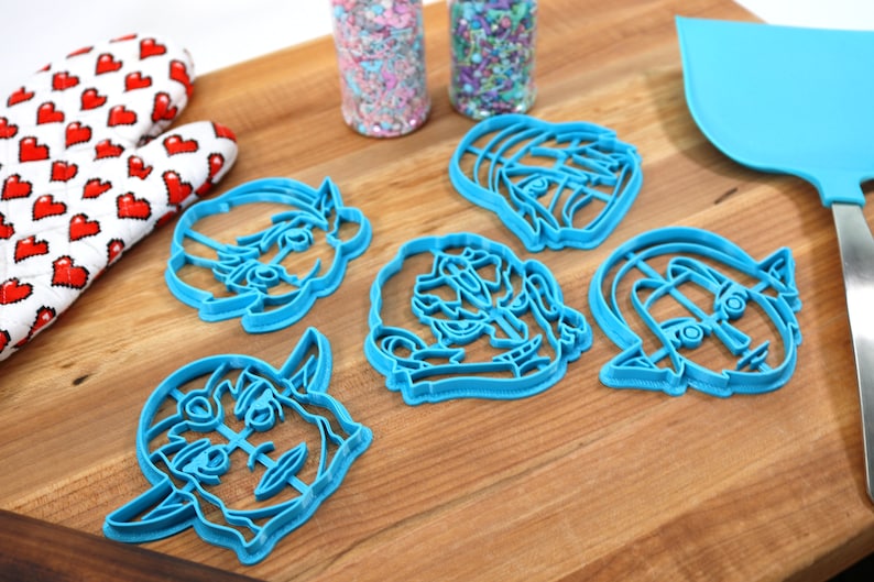 May include: Five blue plastic cookie cutters shaped like cartoon faces. The cutters are on a wooden surface.