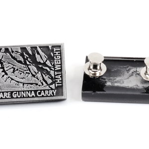 May include: A silver metal pin with a black and white image of a cracked surface. The pin reads "You are gonna carry that weight". The back of the pin is black with two silver metal clasps.