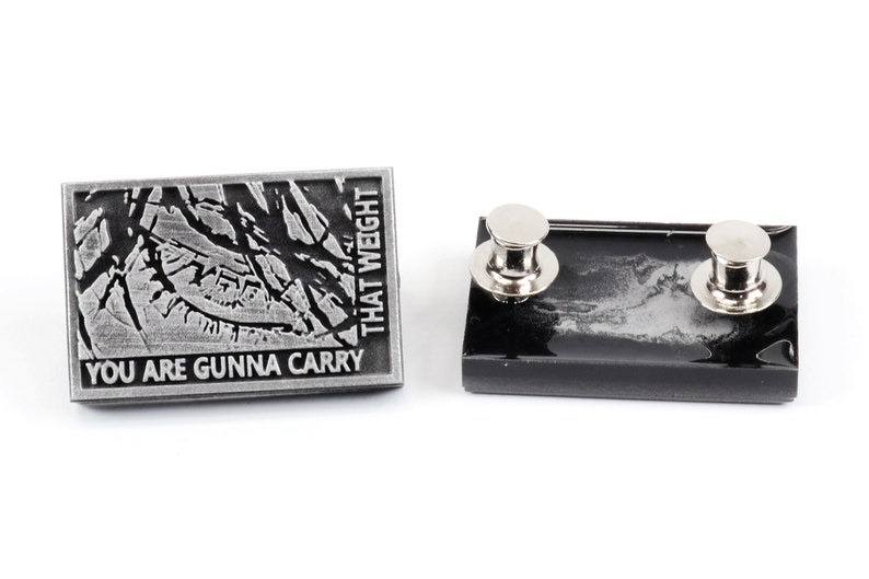 May include: A silver metal pin with a black and white image of a cracked surface. The pin reads "You are gonna carry that weight". The back of the pin is black with two silver metal clasps.
