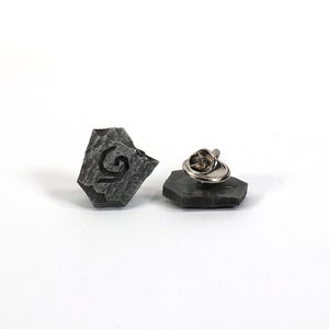 Puede incluir: Un pin de piedra gris con un dise&ntilde;o estilizado de la letra G. El pin tiene un cierre de metal en la parte posterior.