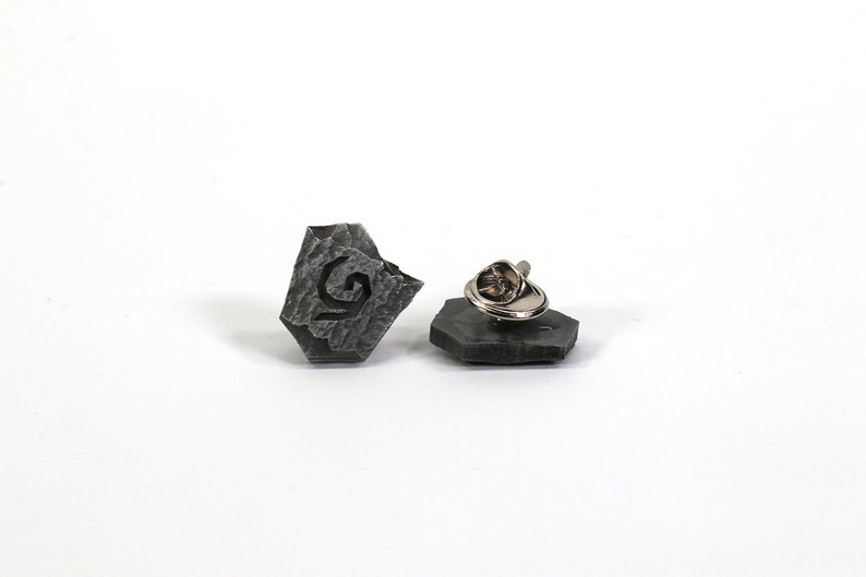 Puede incluir: Un pin de piedra gris con un dise&ntilde;o estilizado de la letra G. El pin tiene un cierre de metal en la parte posterior.