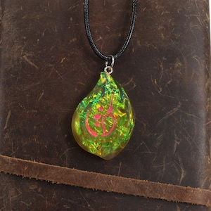 May include: A green and gold iridescent teardrop-shaped pendant with a pink symbol inside, hanging from a black cord necklace. The pendant is resting on a brown leather surface.