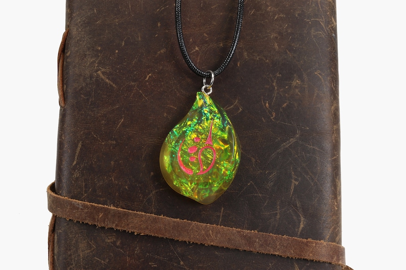May include: A green and gold iridescent teardrop-shaped pendant with a pink symbol inside, hanging from a black cord necklace. The pendant is resting on a brown leather surface.