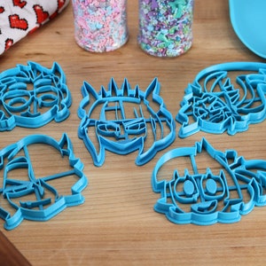 May include: Five blue plastic cookie cutters shaped like cartoon faces. The cutters are on a wooden surface.
