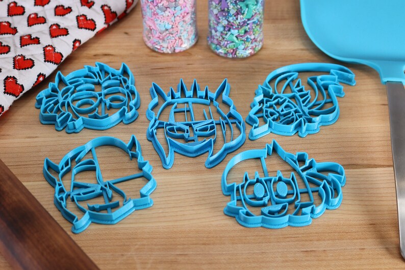 May include: Five blue plastic cookie cutters shaped like cartoon faces. The cutters are on a wooden surface.
