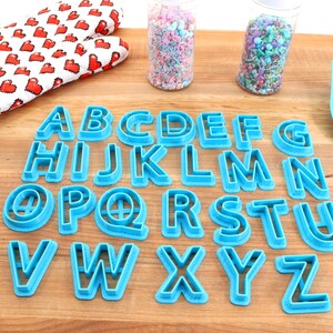 May include: A set of blue plastic alphabet cookie cutters on a wooden surface. The cutters are arranged in alphabetical order. There are two jars of sprinkles in the background.