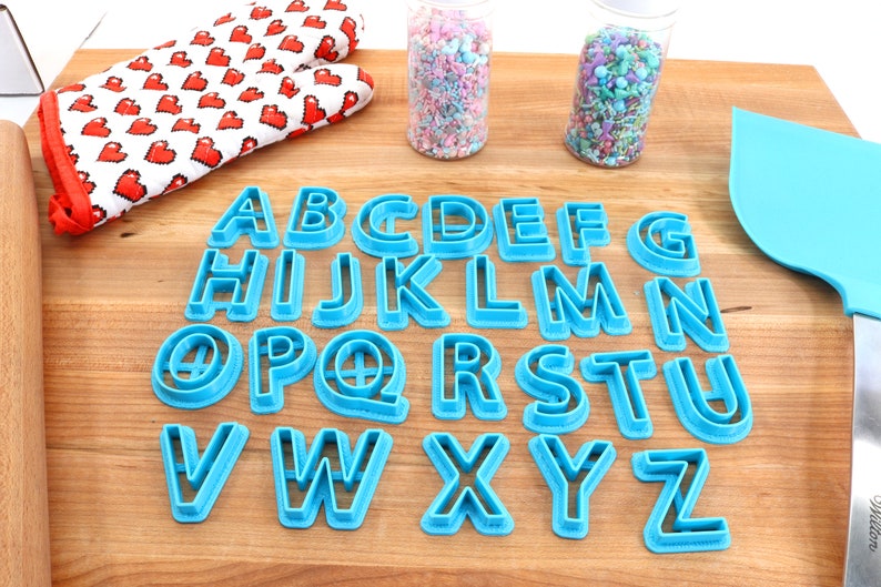 May include: A set of blue plastic alphabet cookie cutters on a wooden surface. The cutters are arranged in alphabetical order. There are two jars of sprinkles in the background.