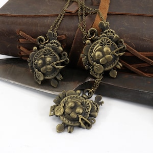 May include: Three bronze steampunk-style necklaces with intricate gears and clockwork designs. The necklaces are hanging from a brown leather journal and a knife.