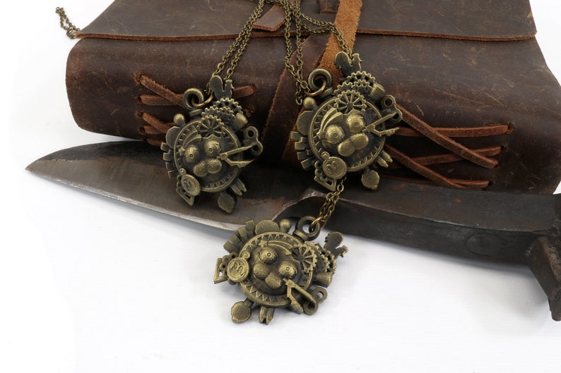 May include: Three bronze steampunk-style necklaces with intricate gears and clockwork designs. The necklaces are hanging from a brown leather journal and a knife.