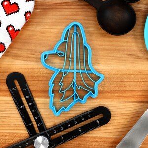 May include: A blue plastic cookie cutter shaped like a dog's head with long hair. The cutter is on a wooden surface with a ruler and a spatula.