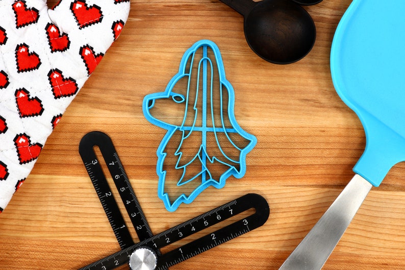 May include: A blue plastic cookie cutter shaped like a dog's head with long hair. The cutter is on a wooden surface with a ruler and a spatula.
