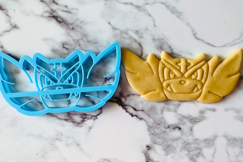 May include: A blue plastic cookie cutter shaped like a cartoon monster with sharp teeth and a menacing expression. Next to it is a cookie baked in the shape of the monster, with a light brown color.